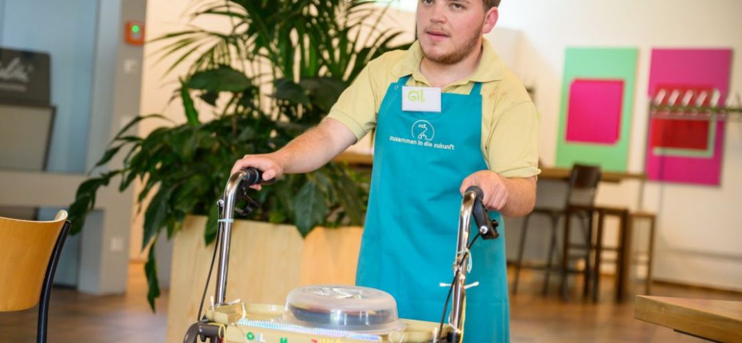 Ein Teilnehmer von “besser zusammen - Berufsorientierung in der Gastronomie für Jugendliche und junge Menschen mit Behinderungen" bringt ein Essen mit seinem Rollwagen an den  Tisch