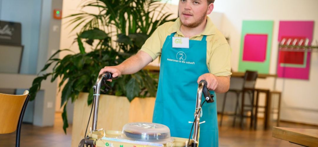Ein Teilnehmer von “besser zusammen - Berufsorientierung in der Gastronomie für Jugendliche und junge Menschen mit Behinderungen" bringt ein Essen mit seinem Rollwagen an den  Tisch