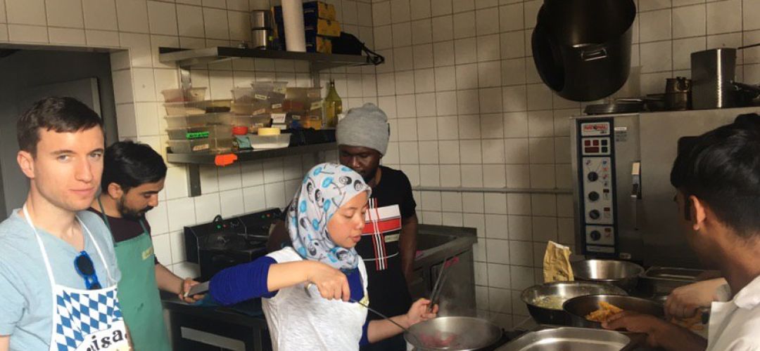 Fünf Personen stehen am Herd bzw. in der Küche und kochen gemeinsam.