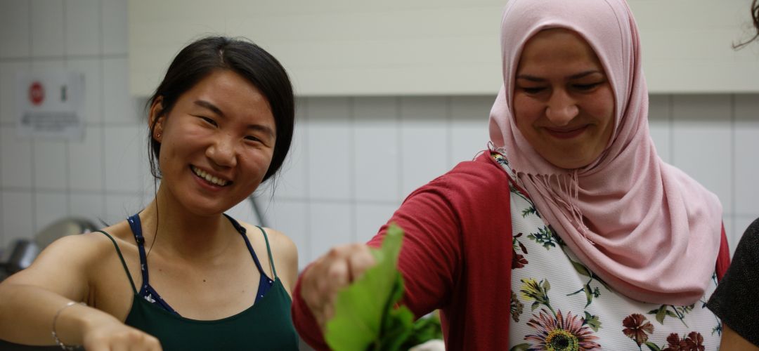 Zwei Workshop Teilnehmer*innen sind in der Küche und geben Zutaten in die Töpfe.