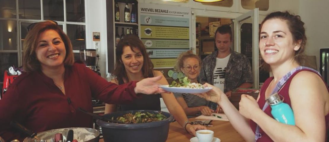 Das Thekenteam zusammen mit der Köchin bereitet den teller zur Ausgabe an eine Gästin vor und überreicht den Teller.