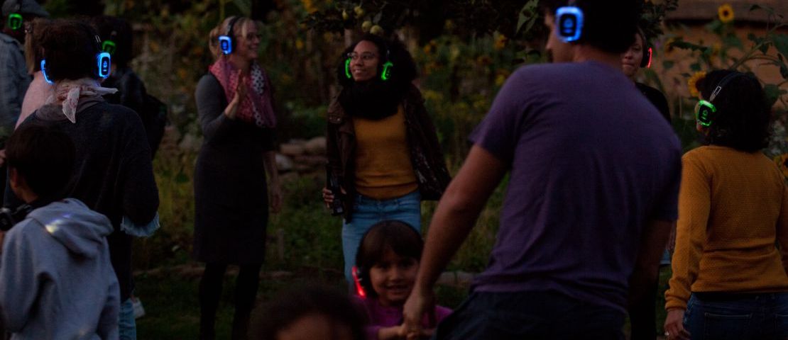 Silent Disco im Gemeinschaftsgarten. Bei Dämmerung tanzen  viele Menschen mit bunten Kopfhörern.