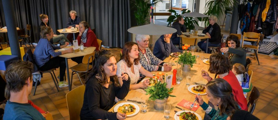 Der Innenraum des zuka solicafé ist zu sehen. Eine Gruppe von 10 Menschen sitzt an einem großen Holztisch in der Mitte, 8 Frauen, ein Mann und ein Kleinkind.  Alle haben Mittagessen vor sich.an den kleineren Tischen sitzen  jeweils 2 Personen.