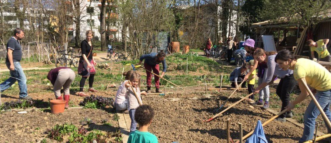 Bei einem AKtionstag im Gemeinschaftsgarten arbeiten  7 Erwachsene und  3 Kinder im großen Gemeinschaftsbeet. Es ist Frühling, der Himmel istblau und die Sonne scheint.