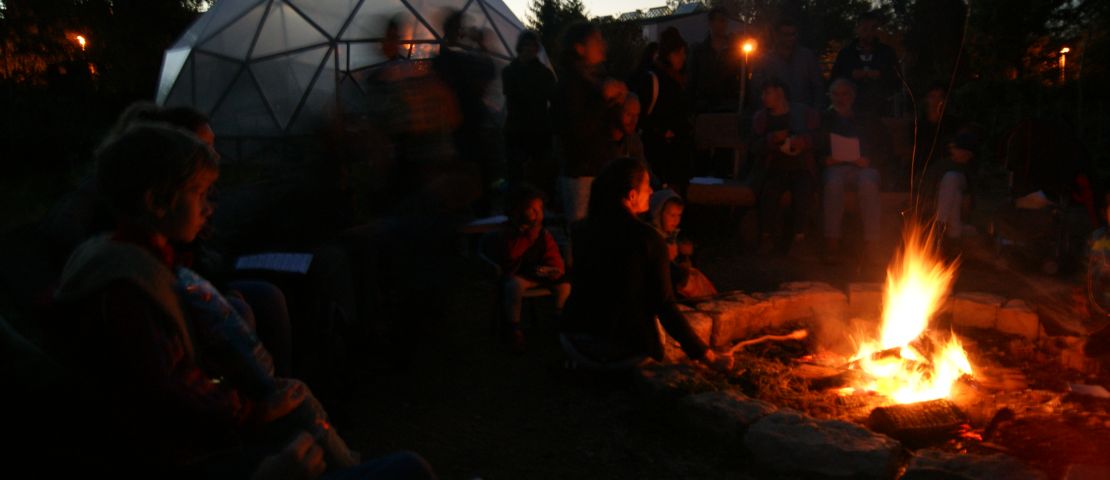 Viele Menschen stehen in der Dämmerung um ein Lagerfeuer im Gemeinschaftsgarten und singen zusammen.