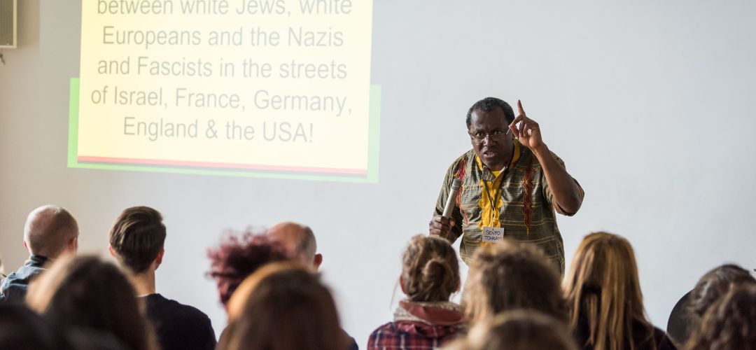 Senfo Tonkam hält einen Vortrag vor Publikum mit Bildschirmpräsentation.