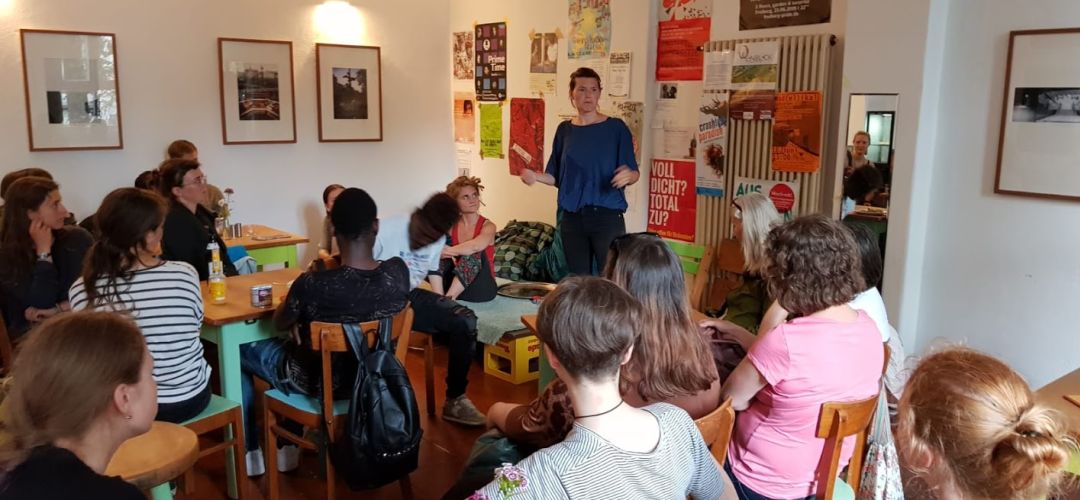 Viele Personen sitzen im Cafe an Tischen und hören einer Person zu die steht und etwas erzählt.