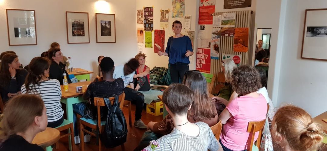 Viele Personen sitzen im Cafe an Tischen und hören einer Person zu die steht und etwas erzählt.