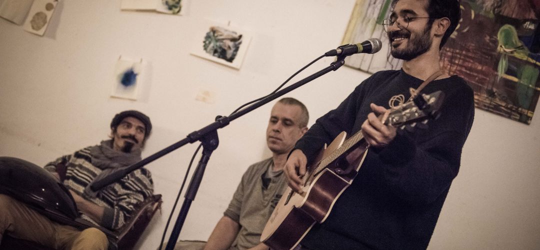 Ein Sänger mit Gitarre am Mikrofon und links davon eine Person and der Cajon und eine Person an der Handdrum.
