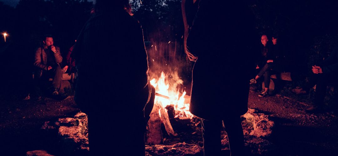 Einige Menschen stehen oder sitzen bei Nacht vor einem Lagerfeuer.
